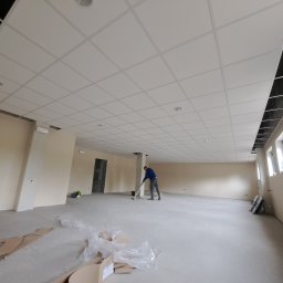 Dawid Kamil - Duże, jasne pomieszczenie z podwieszanym sufitem w trakcie sprzątania po pracach wykończeniowych; widoczny pracownik zamiatający podłogę, resztki materiałów budowlanych i narzędzia przy ścianie.