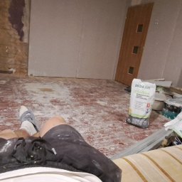 Dawid Kamil - Remontowany pokój z widoczną ścianą z surowej cegły, częściowo zakrytą płytą, workiem z białą gipsową masą szpachlową oraz narzędziami malarskimi i nogami osoby siedzącej na kanapie.