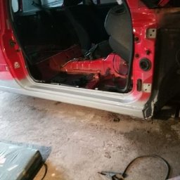 Dawid Kamil - Czerwony samochód osobowy w trakcie naprawy blacharskiej, widoczny próg po nałożeniu szpachli, otwarte drzwi i wnętrze pojazdu z widocznym fragmentem siedzenia, podłoga warsztatu z narzędziami w tle.