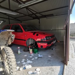 Dawid Kamil - Czerwony samochód z czarnymi naklejkami w kształcie plastra miodu na masce, bez koła, w garażu z motocyklem obok; widoczne narzędzia i resztki naklejek na podłodze.