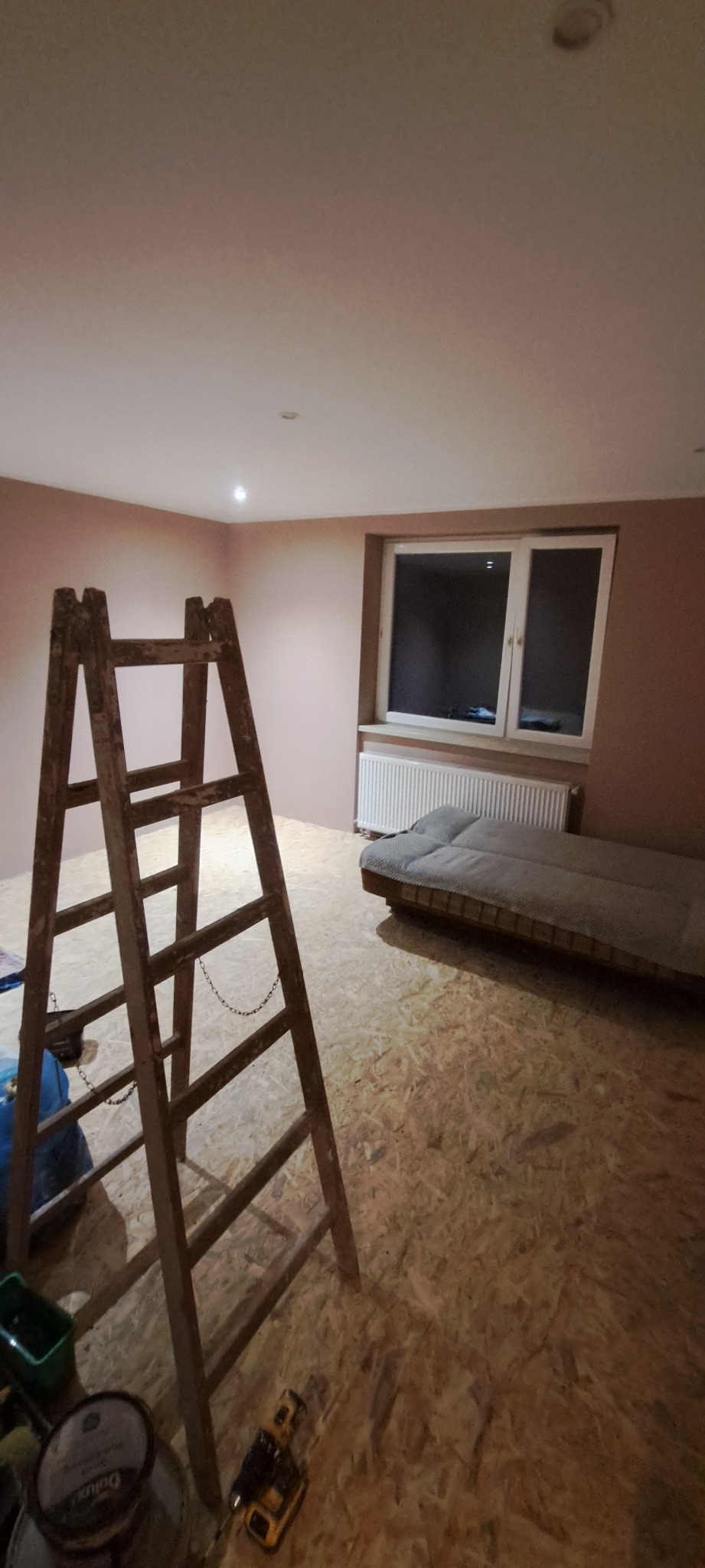 Pokój w trakcie prac wykończeniowych: ściany pomalowane na różowo-brązowy odcień, surowa podłoga z płyt OSB, drewniana drabina, narzędzia i farba na podłodze, widoczne okno z grzejnikiem.