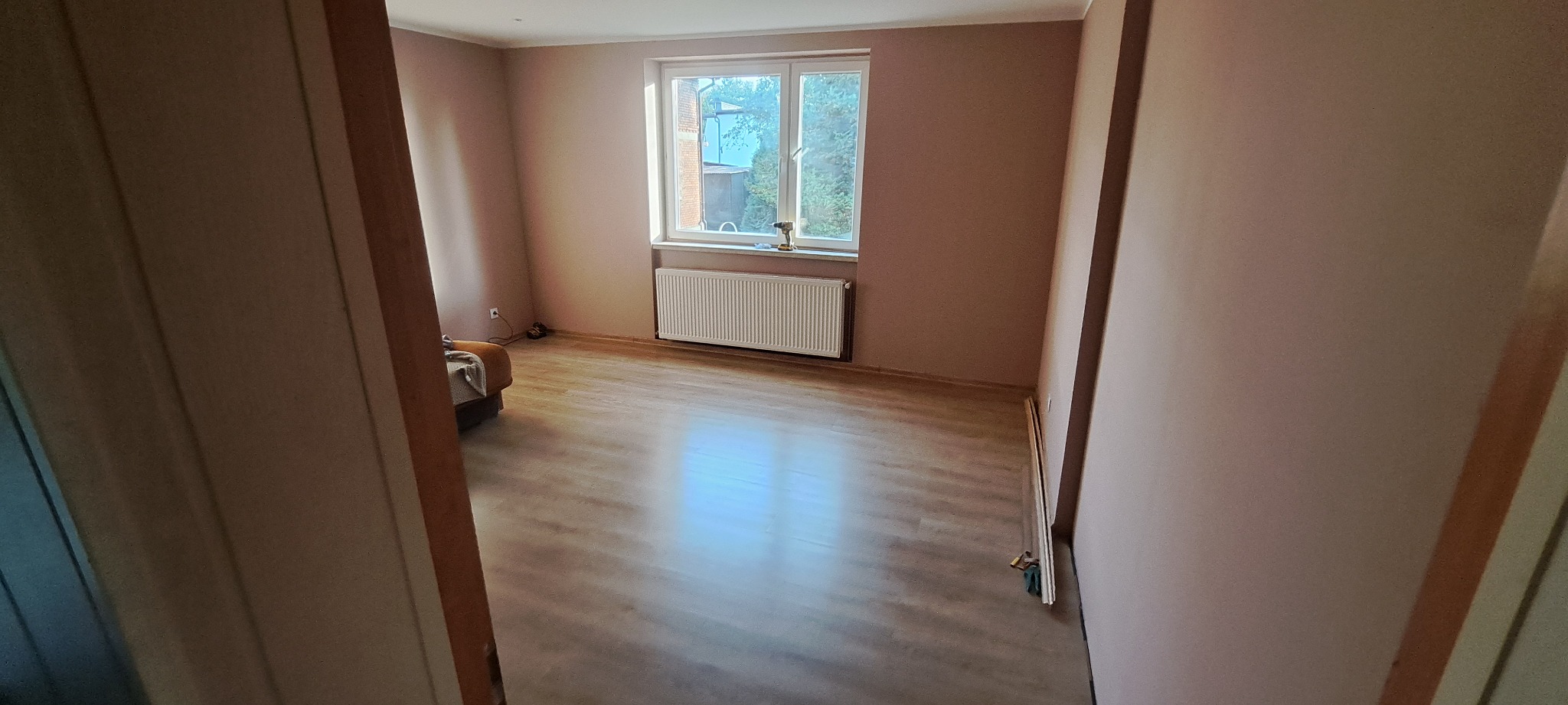 Pokój po malowaniu na kolor pudrowy róż, widoczne panele podłogowe, biały grzejnik pod oknem, częściowo widoczny narożnik i narzędzia przy ścianie.