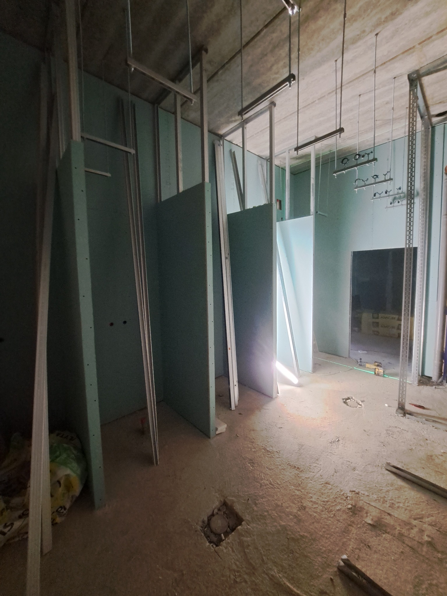 Wnętrze w trakcie montażu ścian działowych z płyt gipsowo-kartonowych na metalowym stelażu. Widoczne niedokończone instalacje pod sufitem i resztki materiałów na podłodze.