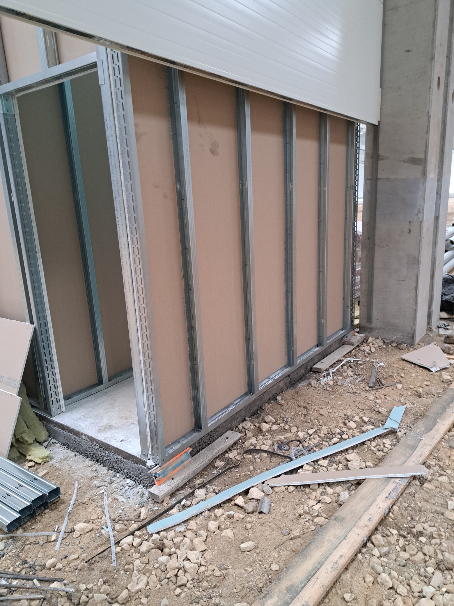 Stelaż ścianki działowej z płyt gipsowo-kartonowych, widoczne profile metalowe i fragmenty płyt, surowy stan wykończenia, betonowy słup w tle.