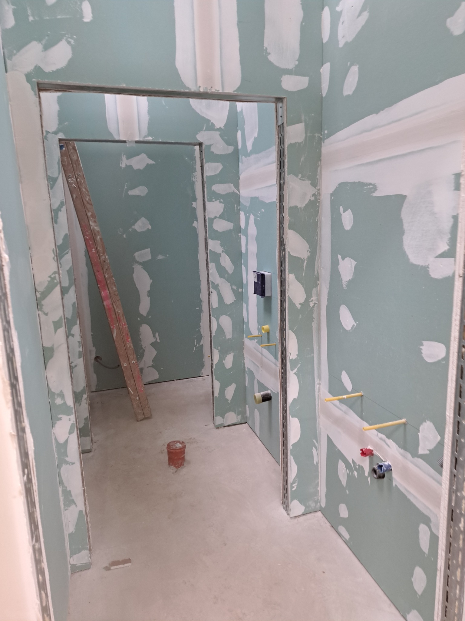 Wnętrze w trakcie remontu: ściany z zielonymi płytami, częściowo pokryte białą masą szpachlową, widoczne profile metalowe, instalacja wodna i drabina oparta o ścianę.