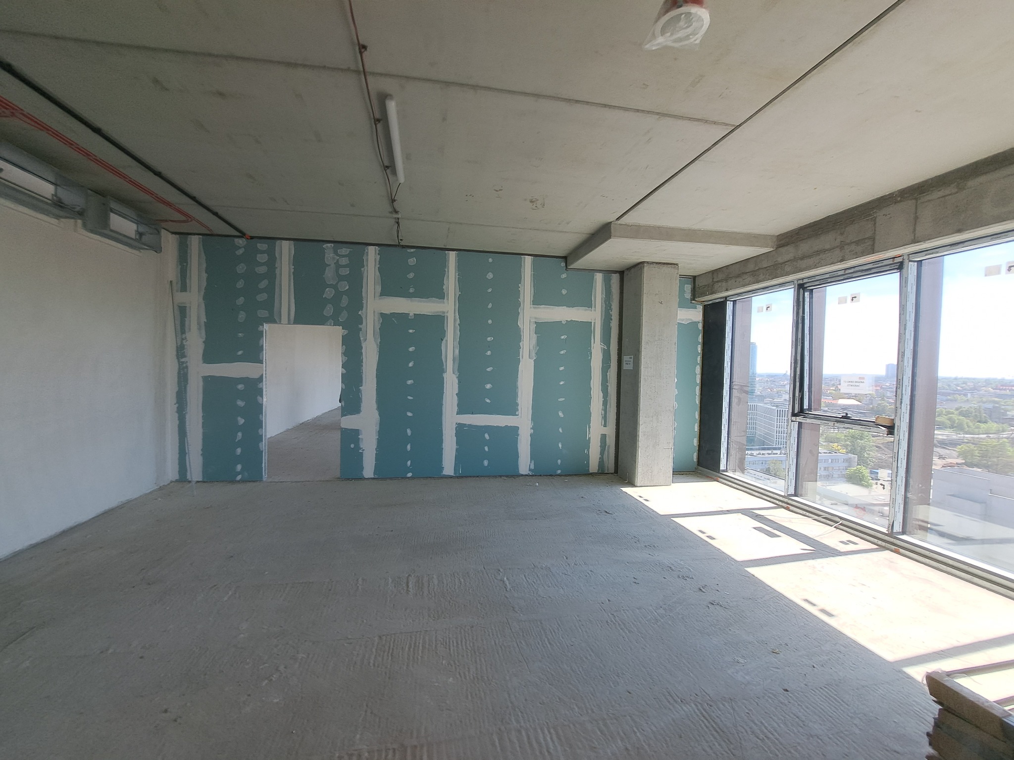 Surowe wnętrze w trakcie prac wykończeniowych, widoczne ściany pokryte niebieskimi płytami z widocznymi łączeniami, surowy beton na podłodze i suficie, duże okna z widokiem na miasto.