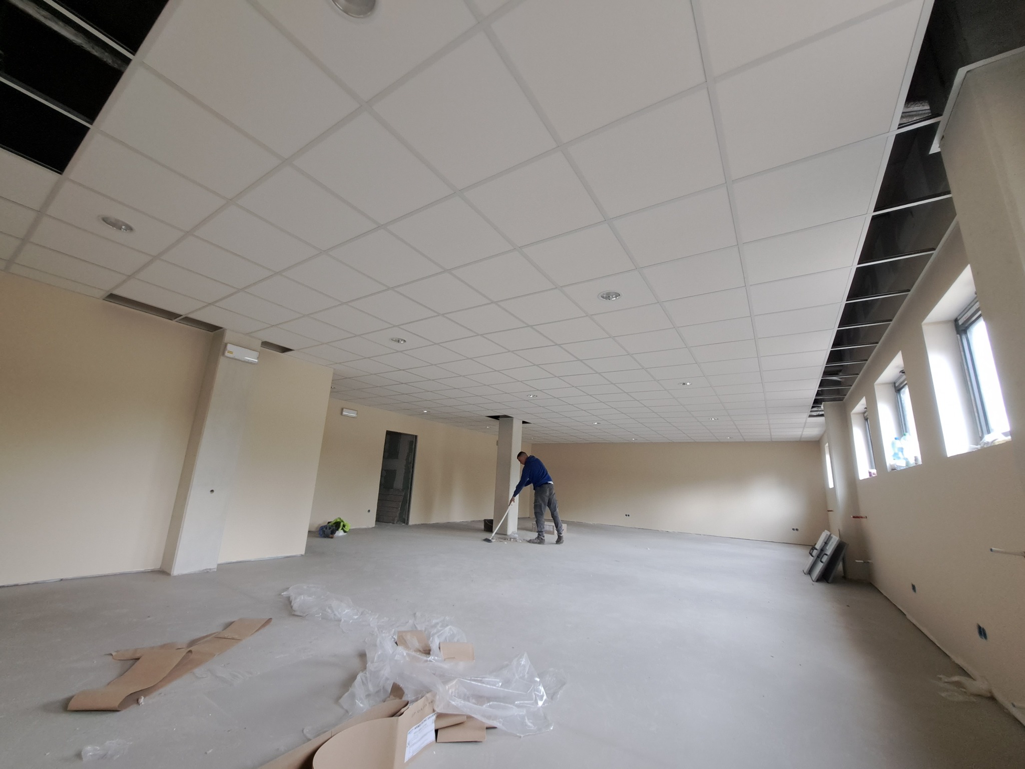 Duże, jasne pomieszczenie z podwieszanym sufitem w trakcie sprzątania po pracach wykończeniowych; widoczny pracownik zamiatający podłogę, resztki materiałów budowlanych i narzędzia przy ścianie.
