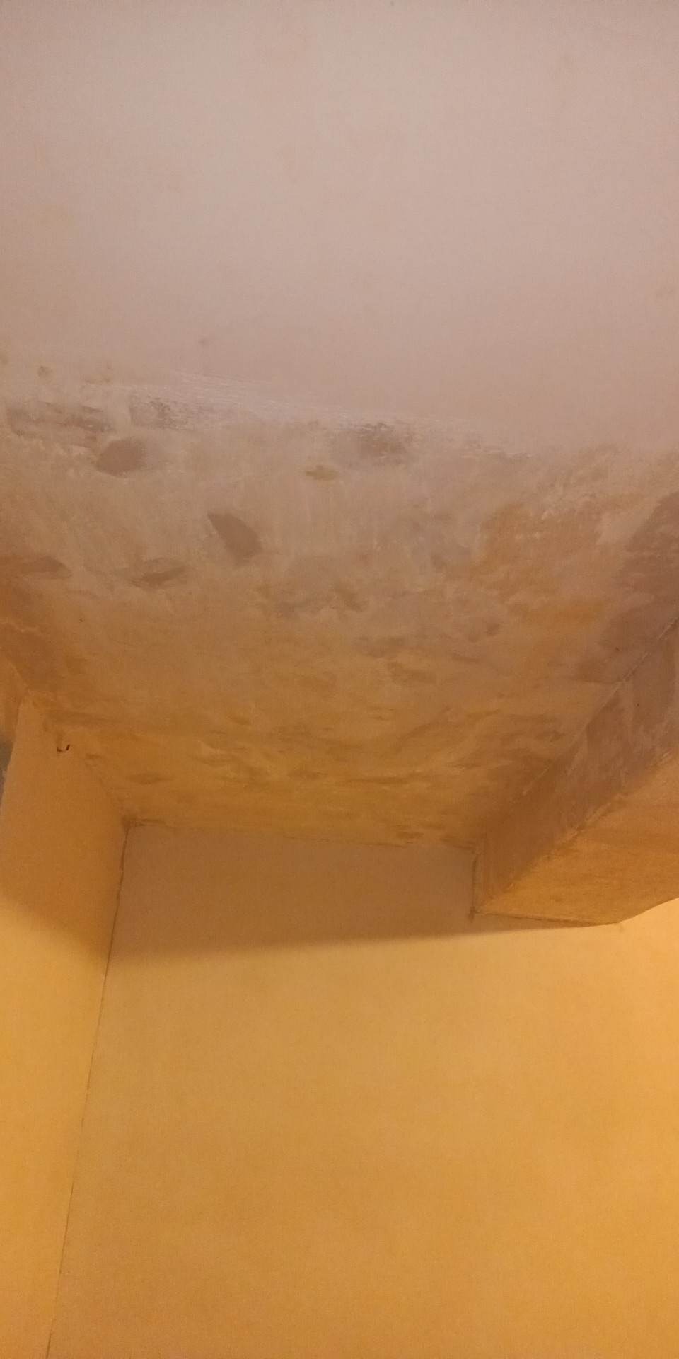 Niedokończone wykończenie sufitu w rogu pomieszczenia, widoczne nierówności i plamy na powierzchni przed nałożeniem ostatecznej warstwy. Ściany pomalowane na żółto.