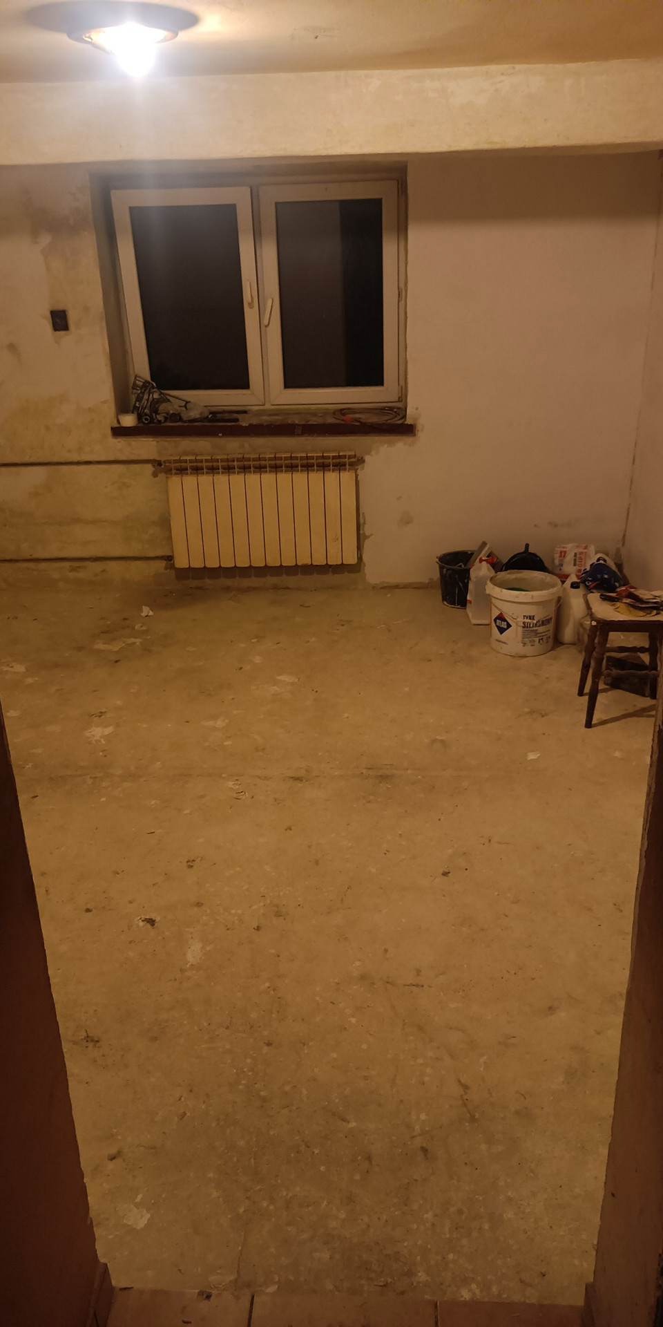 Wnętrze pokoju w trakcie remontu, widoczne surowe ściany, grzejnik, okno, wiadro z masą szpachlową oraz narzędzia malarskie, przygotowane do dalszych prac wykończeniowych.
