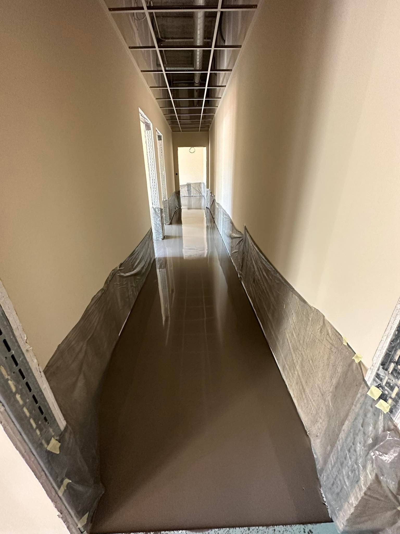Korytarz w trakcie prac wykończeniowych, zabezpieczona podłoga folią, świeżo pomalowane ściany w odcieniach beżu, widoczna surowa konstrukcja sufitu.