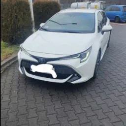 Biała Toyota Corolla kombi zaparkowana na brukowanej nawierzchni, widok z przodu, tablica rejestracyjna zasłonięta, w tle krzewy i niebieski samochód.