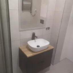 Nowoczesna łazienka z umywalką nablatową na szafce z drewnianym blatem, czarna bateria, grzejnik drabinkowy i lustro z podświetleniem LED na ścianie wyłożonej jasnymi płytkami.