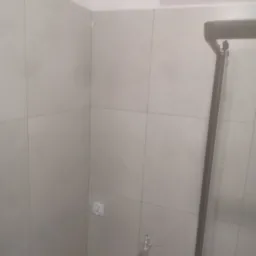 Ściana łazienki wyłożona jasnymi, kwadratowymi płytkami ceramicznymi, z widocznym gniazdkiem elektrycznym i elementami armatury, częściowo zasłonięta ramą kabiny prysznicowej.