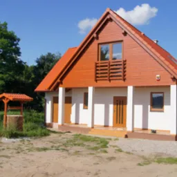 Gotowy, parterowy dom z brązową, drewnianą elewacją i studnią zadaszoną czerwoną dachówką, otoczony zielenią. Ujęcie w słoneczny dzień z niskiej perspektywy.