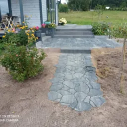 Ścieżka z nieregularnej szarej kostki prowadząca do schodów wejściowych domu. Widoczny fragment trawnika, rabaty z kwiatami i małe drzewko.