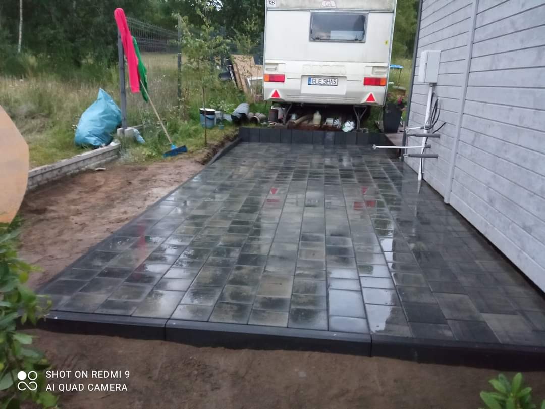 Świeżo ułożona, mokra, szara kostka brukowa z czarnym obrzeżem, widok na zaparkowaną przyczepę kempingową i ścianę budynku gospodarczego.