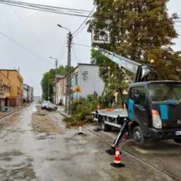 Czarny samochód z podnośnikiem koszowym na mokrej ulicy po wycince gałęzi drzew. W koszu podnośnika widoczna osoba pracująca przy drzewie. Na ulicy leżą połamane gałęzie, a ruch zabezpieczają...