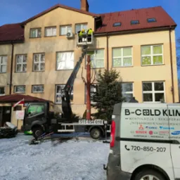 Dwóch pracowników w podnośniku koszowym, w trakcie prac na wysokości przy budynku szkoły, widoczny samochód firmy B-Cold Klima z numerem telefonu i logo.