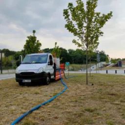 Ciężar&oacute;wka z otwartymi drzwiami, zbiornikiem na wodę i niebieskim wężem podlewa młode drzewo na trawniku w pobliżu drogi.