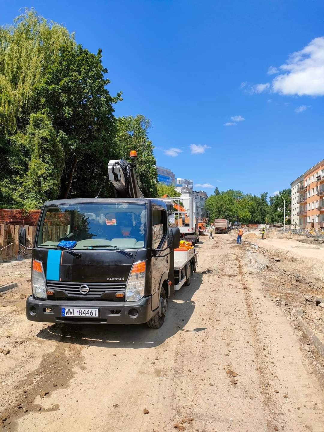 Czarny samochód ciężarowy z wysięgnikiem zaparkowany na placu budowy, w tle drzewa i budynki w słoneczny dzień.
