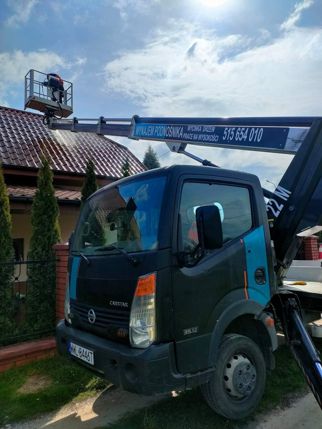 Czarny samochód z podnośnikiem koszowym na tle domu z czerwoną dachówką i błękitnego nieba. Na koszu podnośnika znajduje się osoba w kasku, prawdopodobnie wykonująca prace na wysokości.
