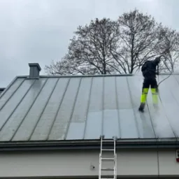 Pracownik w stroju ochronnym i jaskrawych spodniach czyści metalowy dach z zanieczyszczeń za pomocą myjki ciśnieniowej, widoczny fragment dachu przed i po czyszczeniu, drabina oparta o dach...