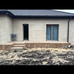 GizoGrod Oliwier Giza - Nowoczesny dom z elewacją z jasnego kamienia i tarasem z kostki. Widoczne schody prowadzące na taras i przygotowany teren pod ogród. Minimalistyczny design.