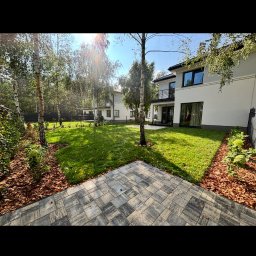 GizoGrod Oliwier Giza - Ułożona kostka brukowa prowadząca do zadbanego trawnika z drzewami i nowoczesnym domem w tle, w słoneczny dzień. Widok na ogród z perspektywy kostki.