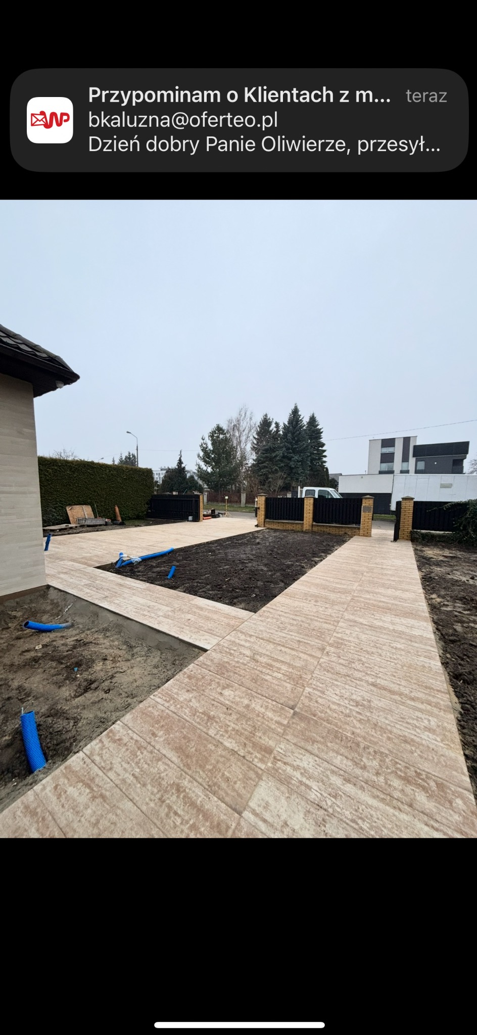 Układanie beżowej kostki brukowej na posesji, widoczne niebieskie rury w wykopie. Częściowo ukończony chodnik prowadzi do bramy wjazdowej z czarnym ogrodzeniem.