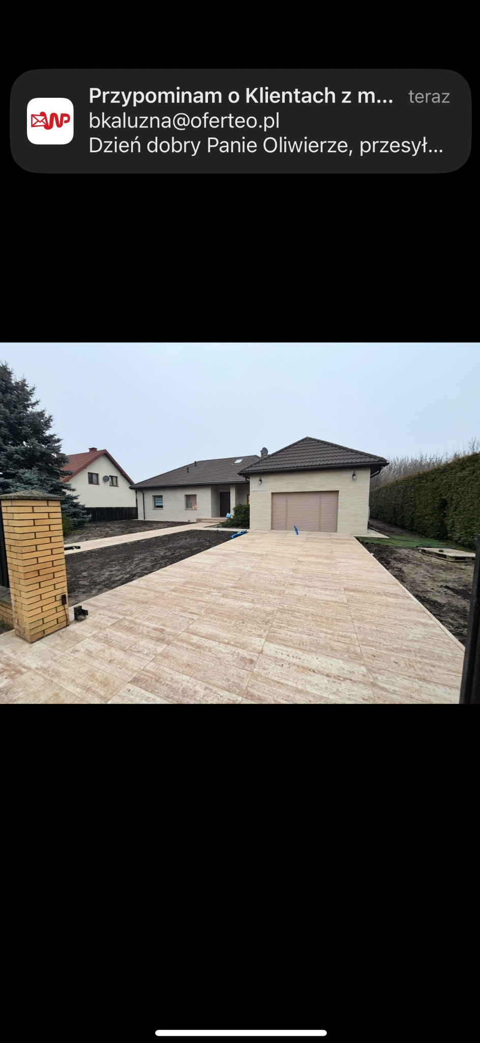 Ułożona kostka brukowa na podjeździe do domu z garażem, widok z perspektywy ogrodu. Świeżo obsiana ziemia po bokach. Dom z brązowym dachem w tle.