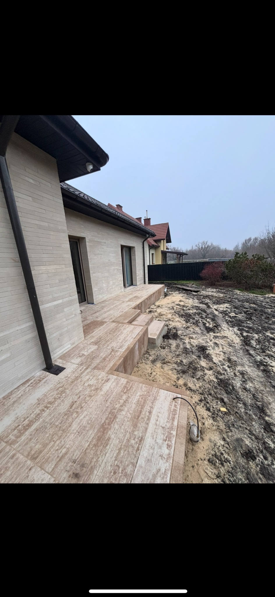 Wykończony taras z płytek imitujących drewno przy domu z elewacją w jasnym odcieniu. Widoczne schody tarasowe i przygotowany teren pod ogród.