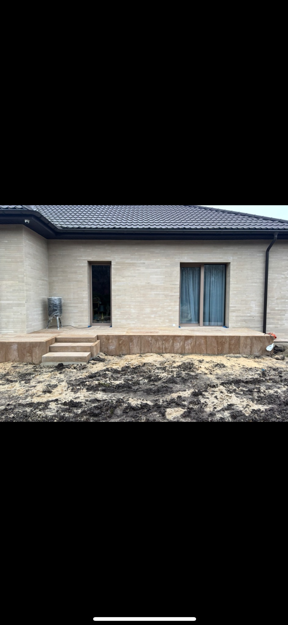 Nowoczesny dom z elewacją z jasnego kamienia i tarasem z kostki. Widoczne schody prowadzące na taras i przygotowany teren pod ogród. Minimalistyczny design.