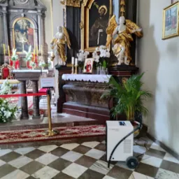 Biały osuszacz powietrza Trotec na kółkach stoi w rogu kościoła z marmurową posadzką w szachownicę, w tle ołtarz z figurami aniołów.