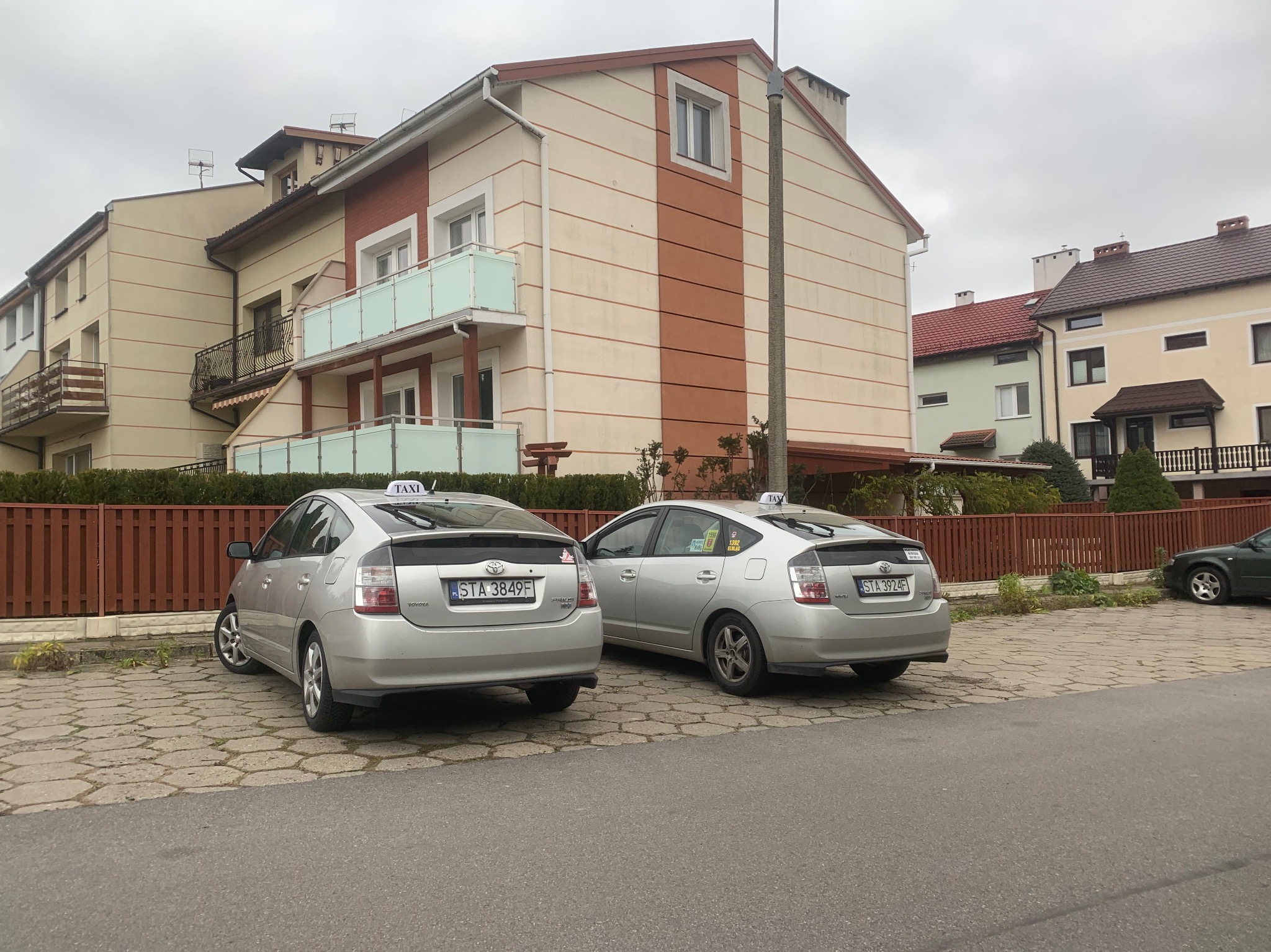 Dwa srebrne samochody Toyota Prius z oznaczeniami Taxi stoją zaparkowane na kostce brukowej przed budynkiem mieszkalnym w pochmurny dzień.