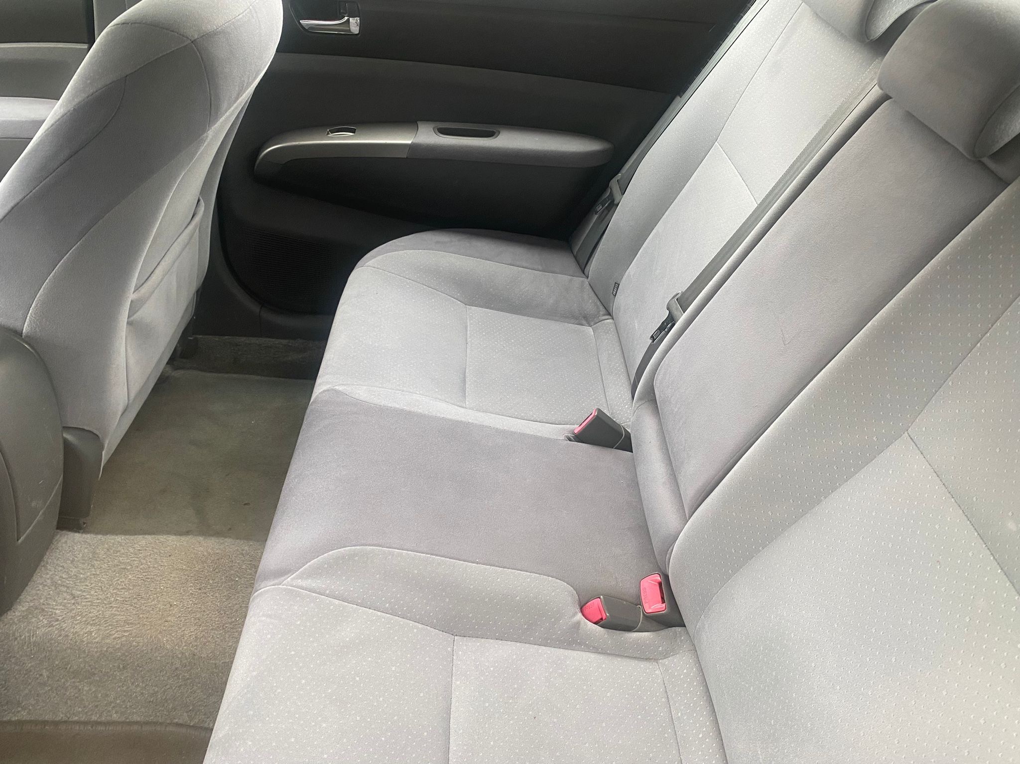 Wnętrze samochodu z tylnymi siedzeniami z szarej tapicerki z widocznymi pasami bezpieczeństwa z różowymi elementami, uchwytem i panelem sterowania szybą w drzwiach.