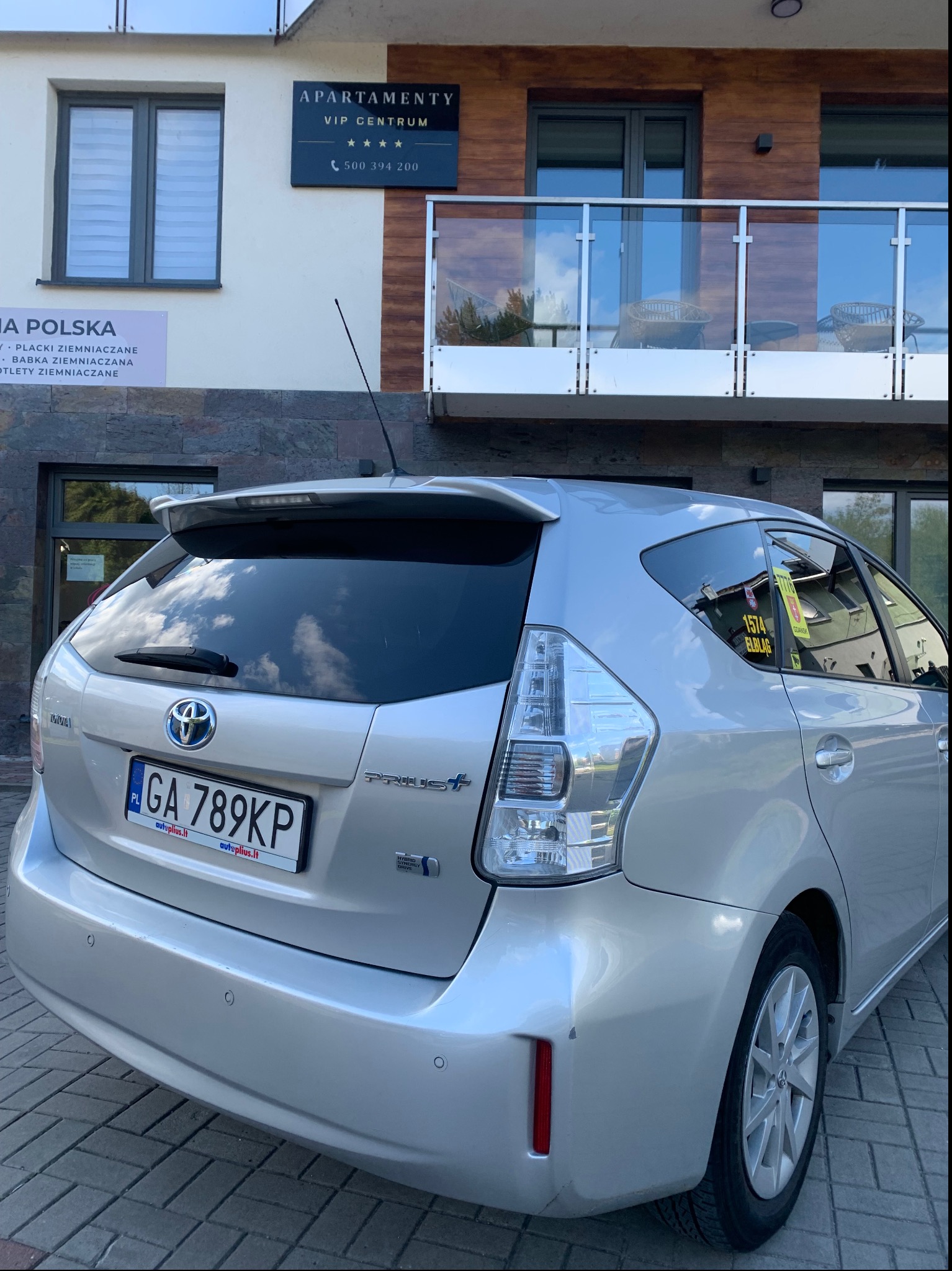 Srebrny Toyota Prius+ z naklejką '1574 Elbląg' na tle budynku 'Apartamenty VIP Centrum'. Tablica rejestracyjna GA 789KP. Widok z tyłu, lekki kąt.