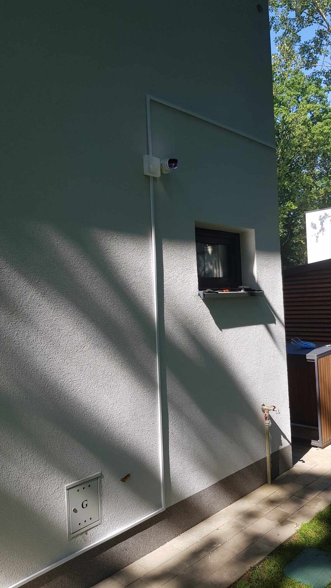 Zewnętrzna kamera bezpieczeństwa zamontowana na elewacji domu, z widocznymi przewodami w białych listwach, oświetlona promieniami słońca.