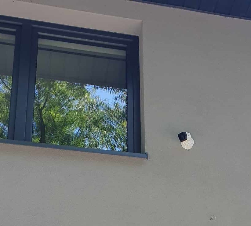 Kamera IP zamontowana na elewacji domu obok okna z odbiciem drzewa.