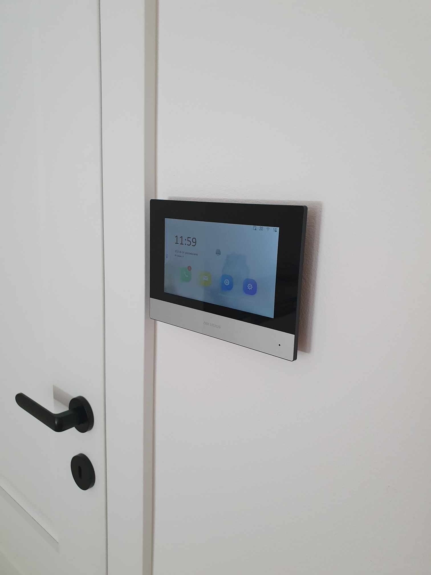 Panel dotykowy wideodomofonu marki Hikvision zamontowany na białej ścianie obok białych drzwi z czarną klamką.