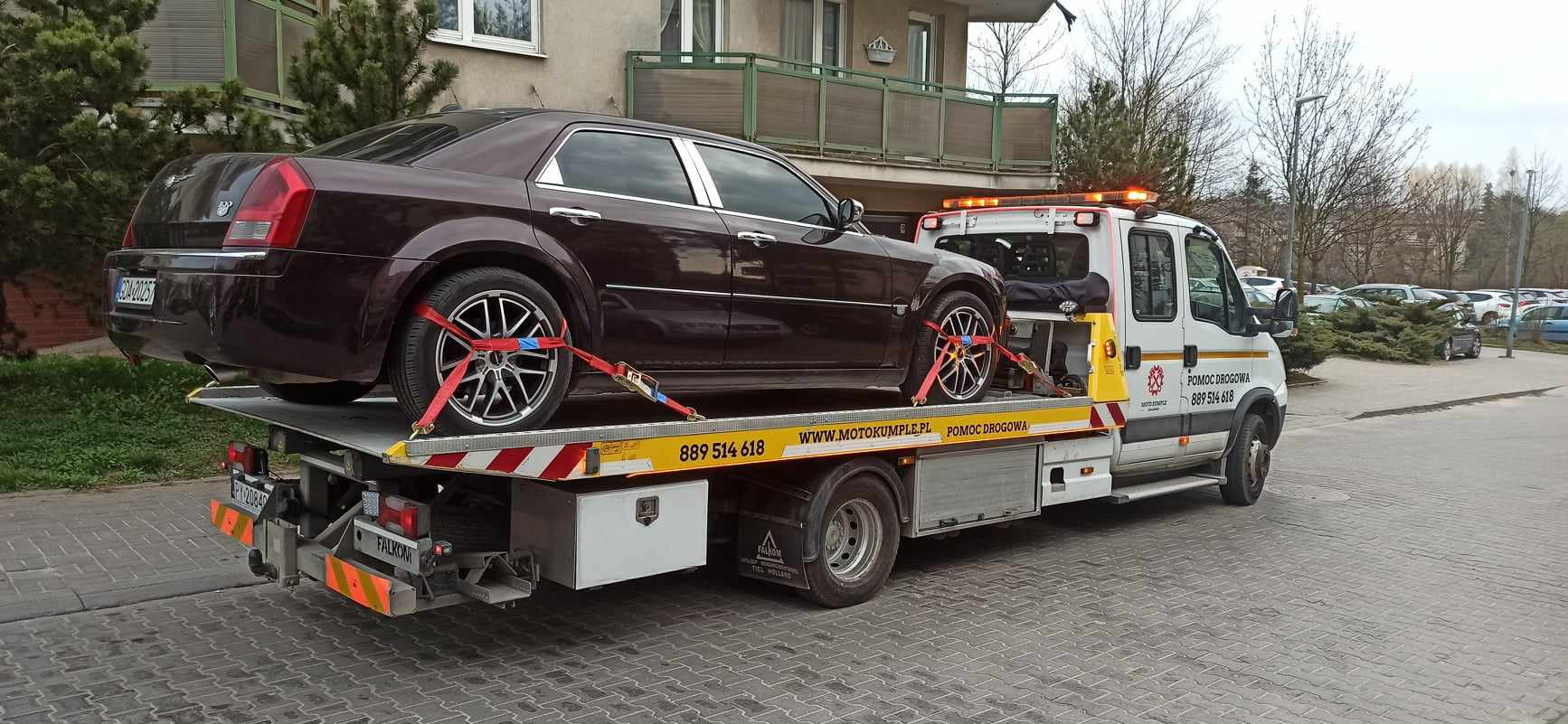 Bordowy samochód osobowy, zabezpieczony czerwonymi pasami transportowymi, przewożony na platformie pojazdu pomocy drogowej z włączonymi światłami ostrzegawczymi na tle budynku mieszkalnego i parkingu.