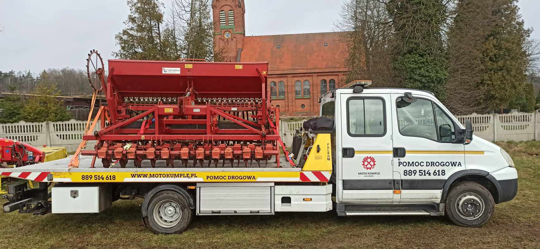 Biała ciężarówka pomocy drogowej z czerwonym siewnikiem rolniczym na platformie, widoczne logo firmy i numer telefonu, w tle ceglany budynek z wieżą zegarową.