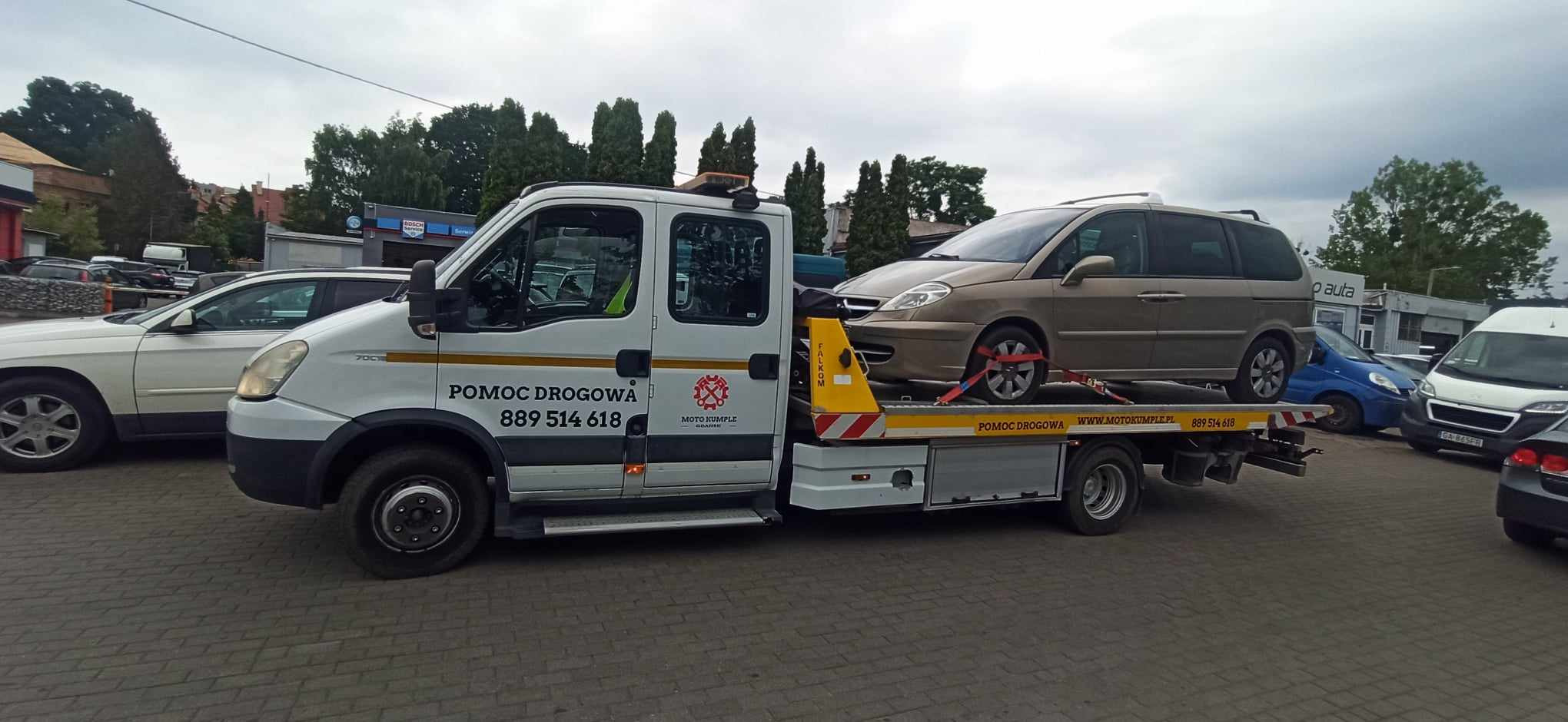 Laweta Moto Kumple z Gdańska przewozi beżowego minivana zabezpieczonego czerwonymi pasami transportowymi na tle budynków.