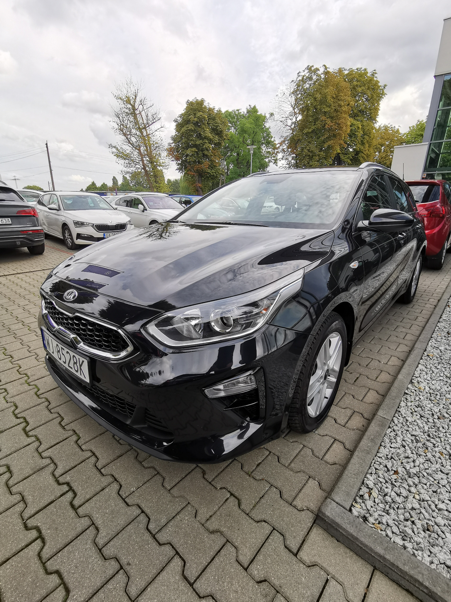 Czarny samochód Kia Ceed kombi, zaparkowany na kostce brukowej z widoczną tablicą rejestracyjną, na tle innych aut i budynku z przeszkloną fasadą.