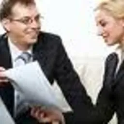 Mężczyzna w okularach, prawdopodobnie doradca, prezentuje dokument kobiecie w garniturze; oboje pochyleni nad laptopem, sugerując omówienie wniosku lub biznesplanu.