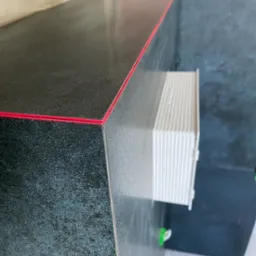 Fragment wykończonej powierzchni z ciemnej, błyszczącej płyty z czerwoną fugą, widoczny fragment białego wentylatora i przewód elektryczny.