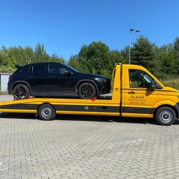 Alkar Pomoc Drogowa - Żółta laweta firmy ALKAR z czarnym samochodem osobowym na platformie, zaparkowana na placu z kostki brukowej w słoneczny dzień.