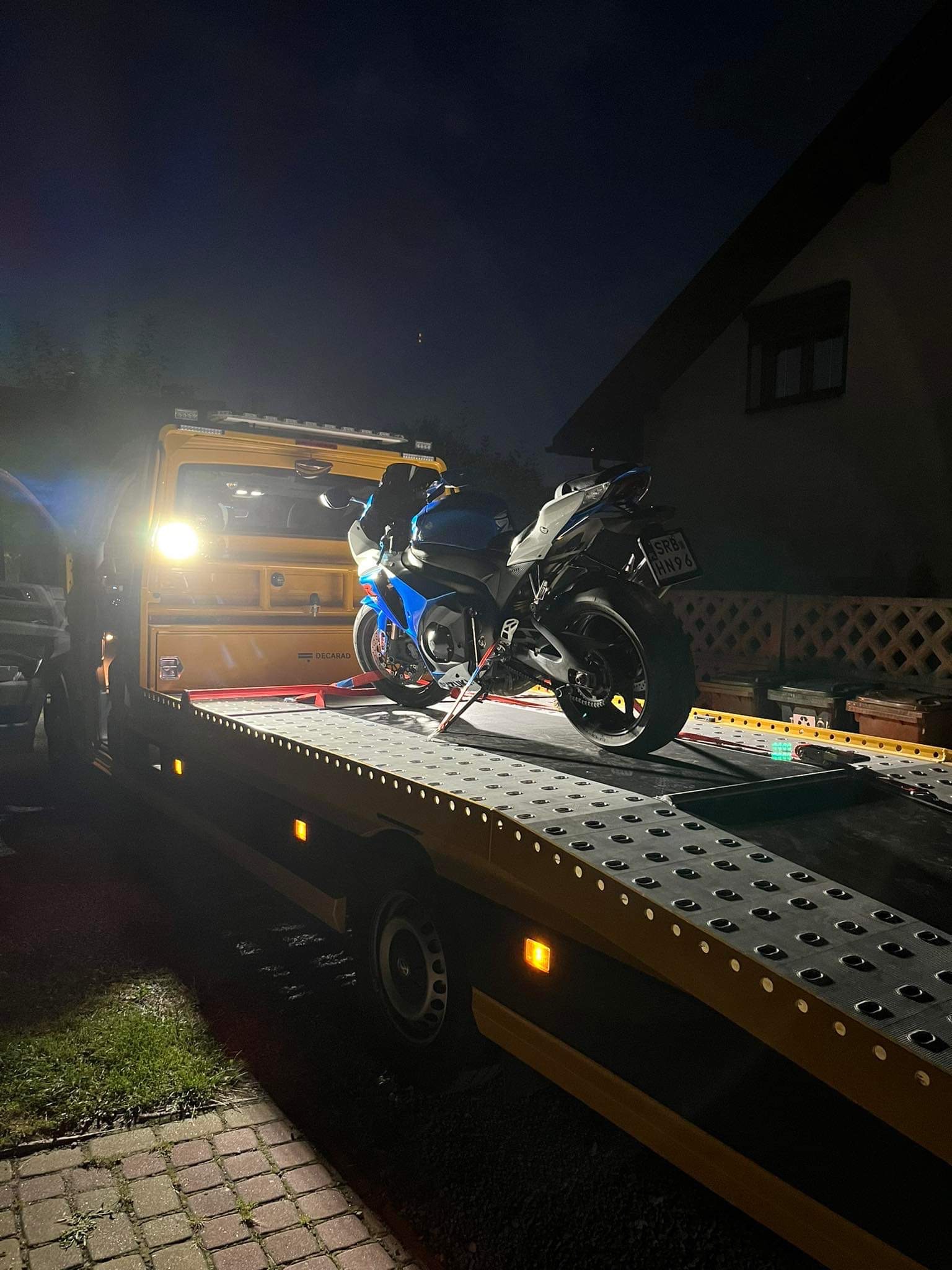 Niebieski motocykl sportowy zabezpieczony pasami na platformie lawety w nocy, widoczne tablice rejestracyjne.