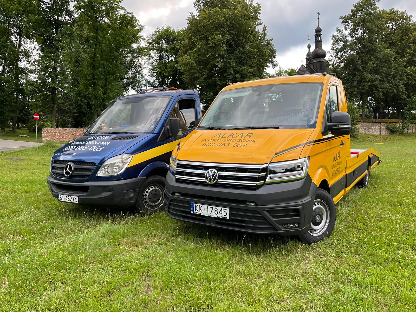 Dwa pojazdy pomocy drogowej: niebieski bus Mercedes i żółta laweta Volkswagen, stoją na zielonej trawie, w tle drzewa i wieża kościoła.
