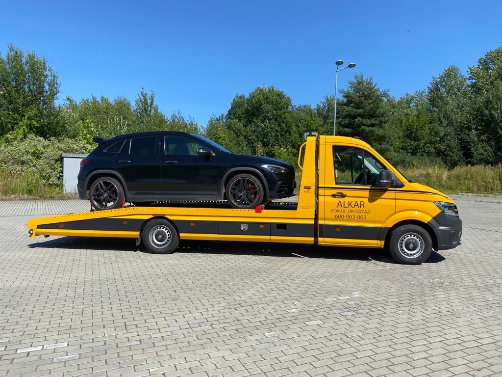 Żółta laweta firmy ALKAR z czarnym samochodem osobowym na platformie, zaparkowana na placu z kostki brukowej w słoneczny dzień.