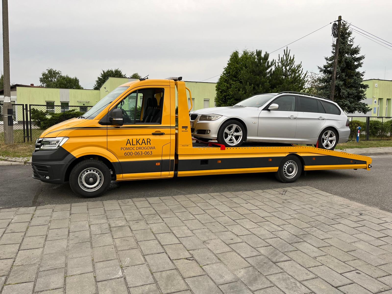 Żółta laweta ALKAR z srebrnym kombi BMW na platformie, zaparkowana na brukowanej ulicy w pochmurny dzień.